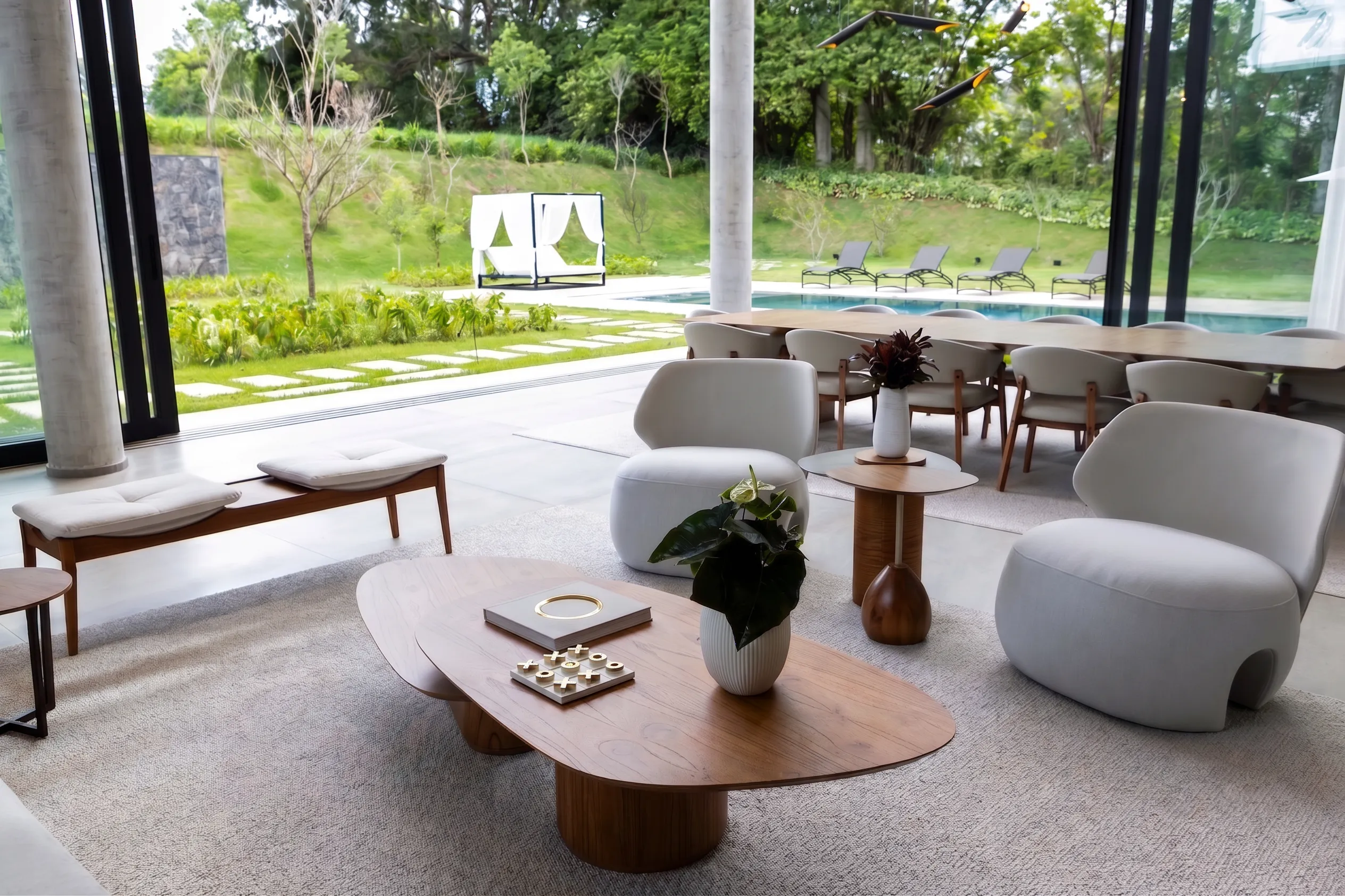 Estar da Casa Entreverdes aberto para piscina e bosque, com mesas orgânicas em madeira, poltronas claras e luz natural