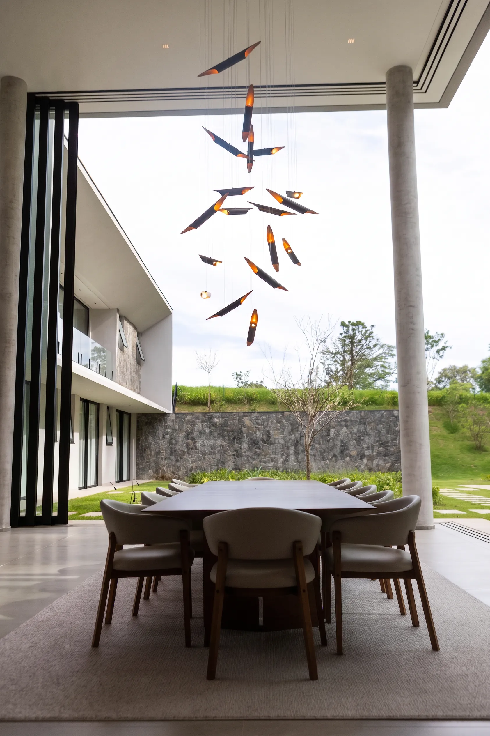 Jantar da Casa Entreverdes na varanda de pé-direito duplo, com pendente escultórico, mesa de madeira e vista para a piscina
