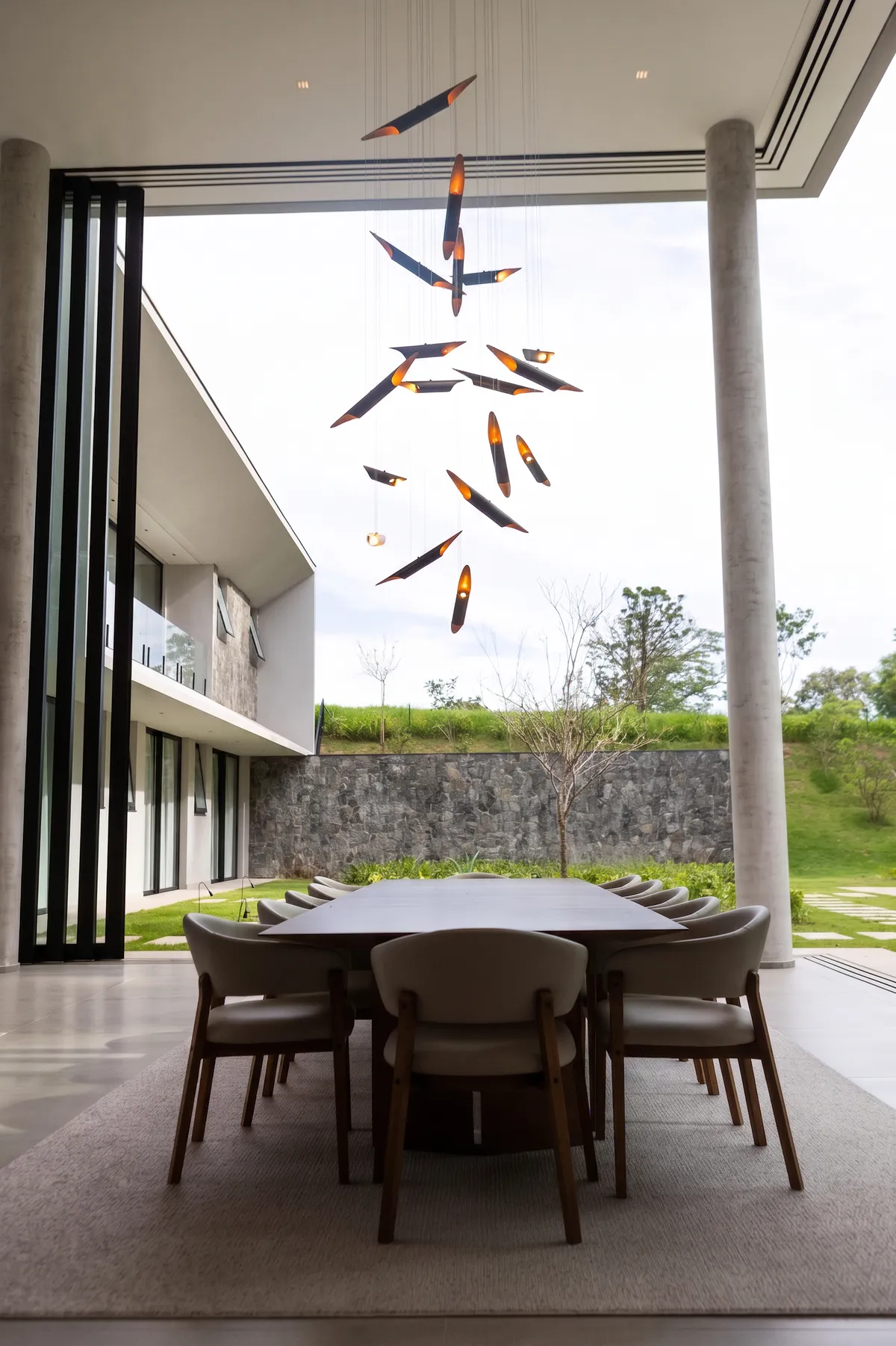 Jantar da Casa Entreverdes na varanda de pé-direito duplo, com pendente escultórico, mesa de madeira e vista para a piscina