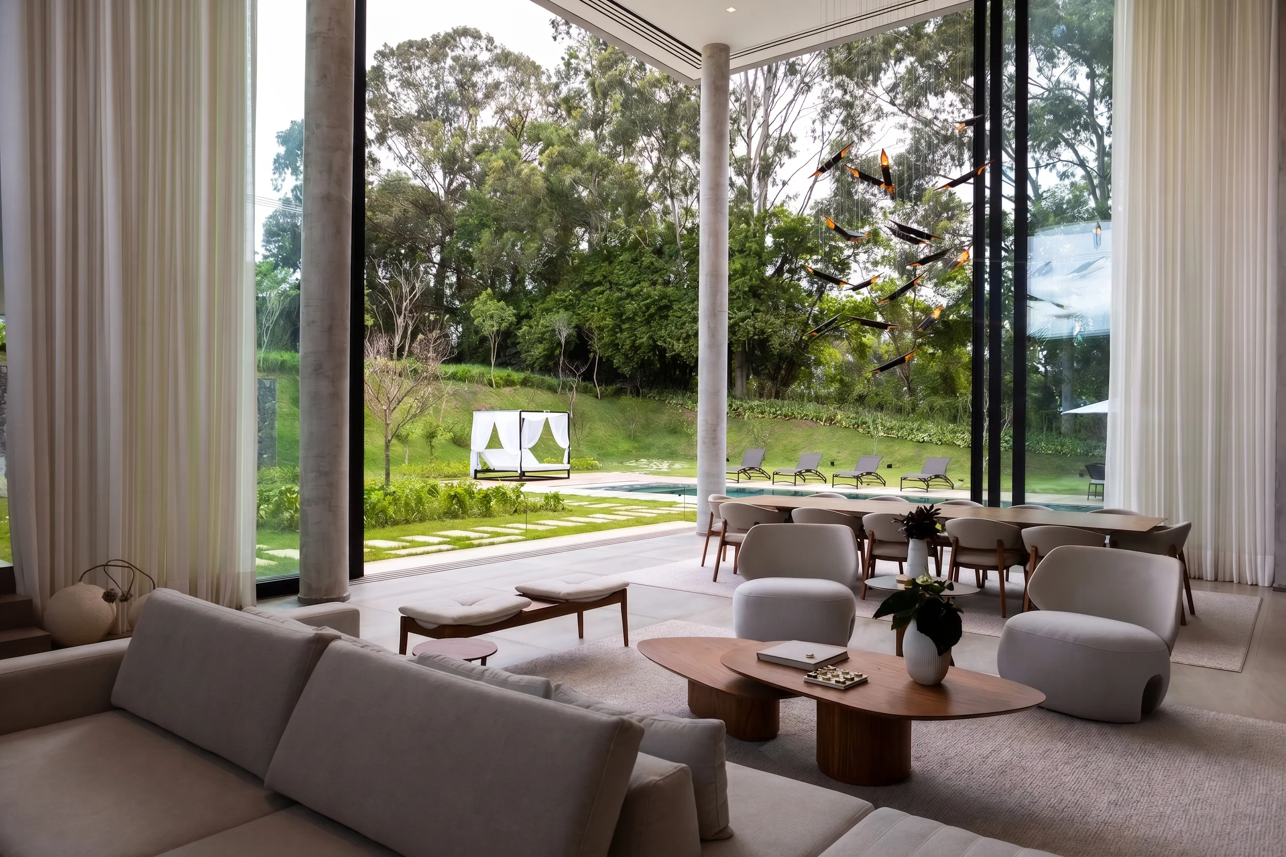 Living da Casa Entreverdes com sofá curvo claro, poltronas, mesas orgânicas, cortinas altas e vista para piscina e bosque