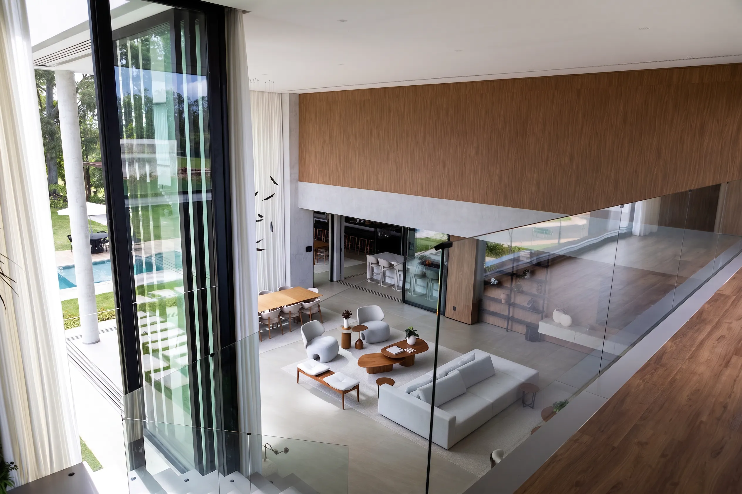Vista superior do living da Casa Entreverdes com pé-direito duplo, painel de madeira, sofá claro e grandes panos de vidro