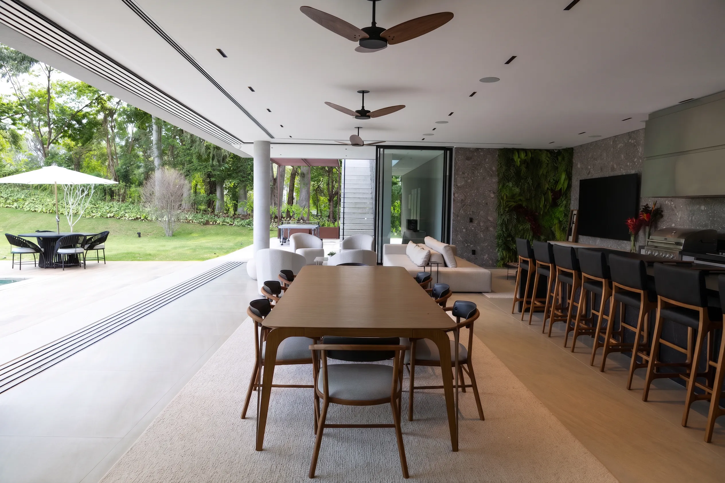 Mesa de jantar da Casa Entreverdes integrada ao gourmet, com bancada em pedra escura, cadeiras de madeira e ventiladores de teto