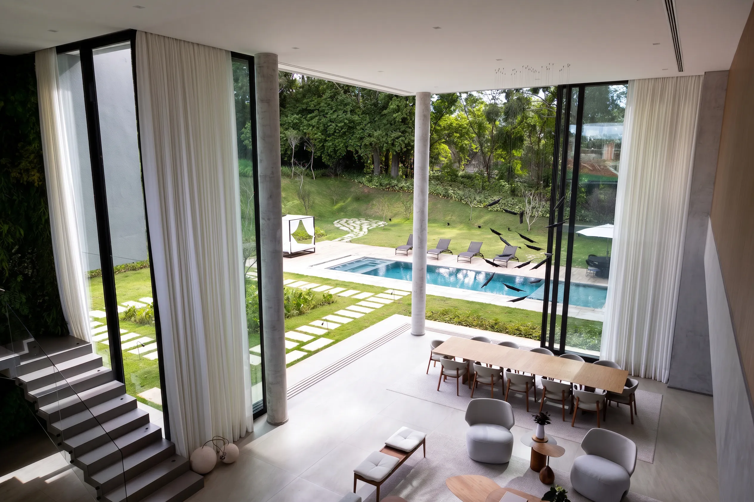 Vista superior da sala integrada da Casa Entreverdes com escada, living, jantar, cortinas altas, piscina e paisagismo externo