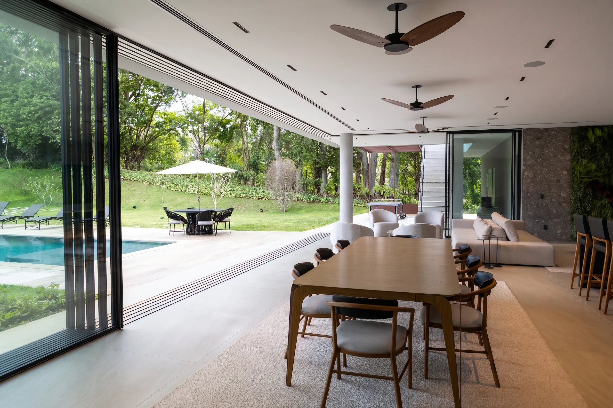 Sala de jantar e varanda gourmet da Casa Entreverdes integradas à piscina, com mesa de madeira, cadeiras estofadas e esquadrias abertas