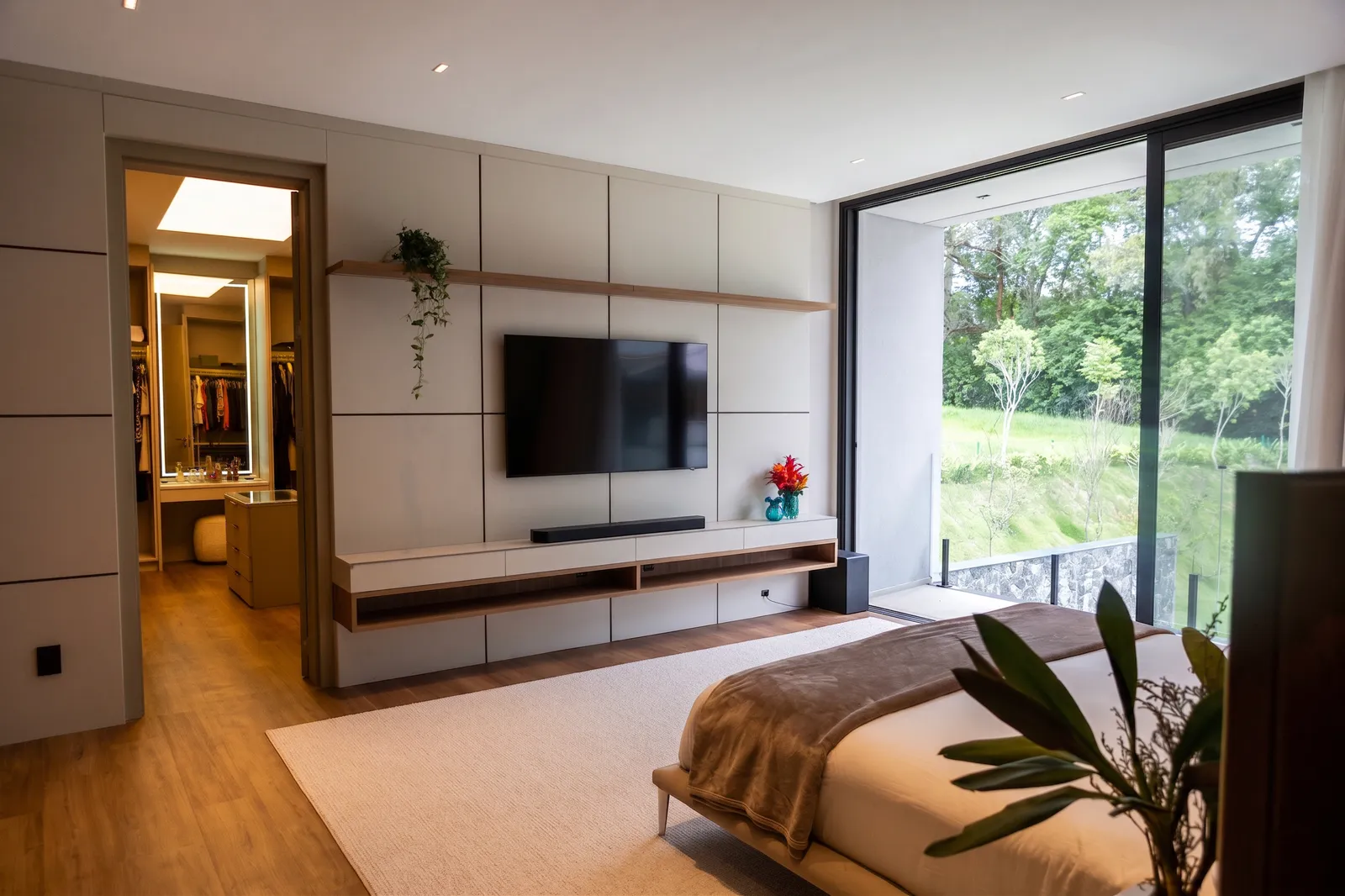 Suíte master da Casa Entreverdes com painel de TV claro, closet iluminado, piso de madeira, cama estofada e vista para o bosque