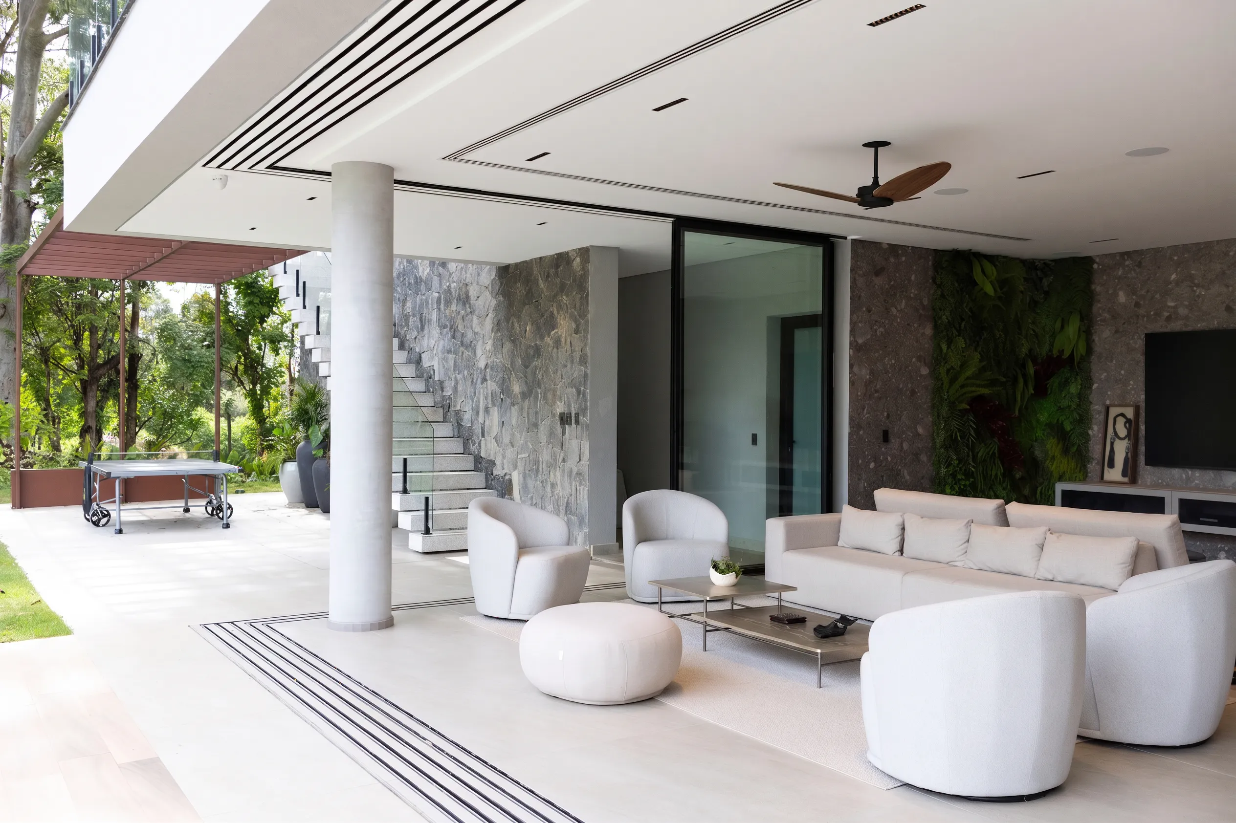 Varanda gourmet da Casa Entreverdes com estar externo, escada em pedra, pilares de concreto, sofá claro e integração com jardim