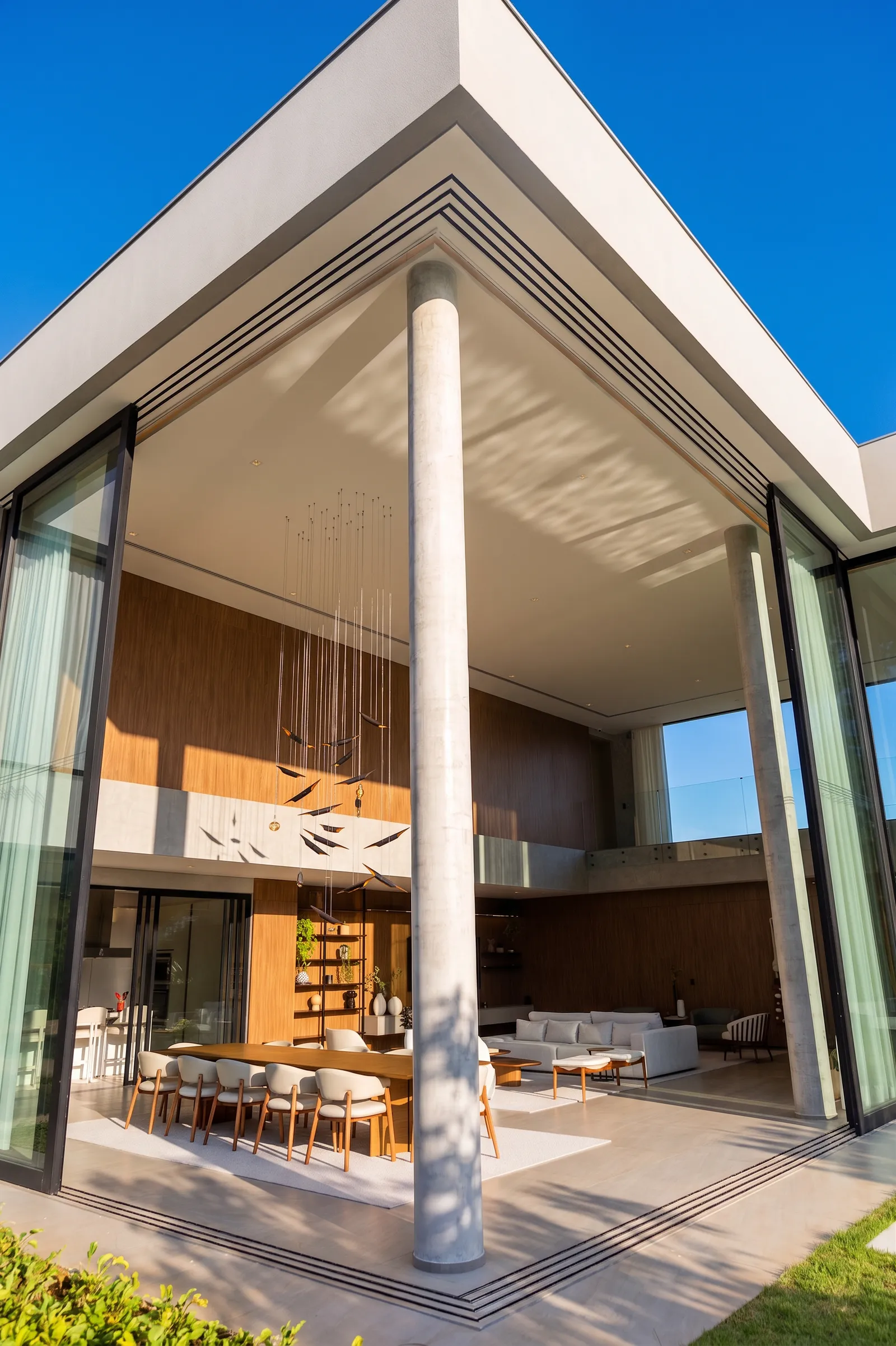Varanda de pé-direito duplo da Casa Entreverdes com pilares cilíndricos, esquadrias de vidro, madeira interna e jardim integrado