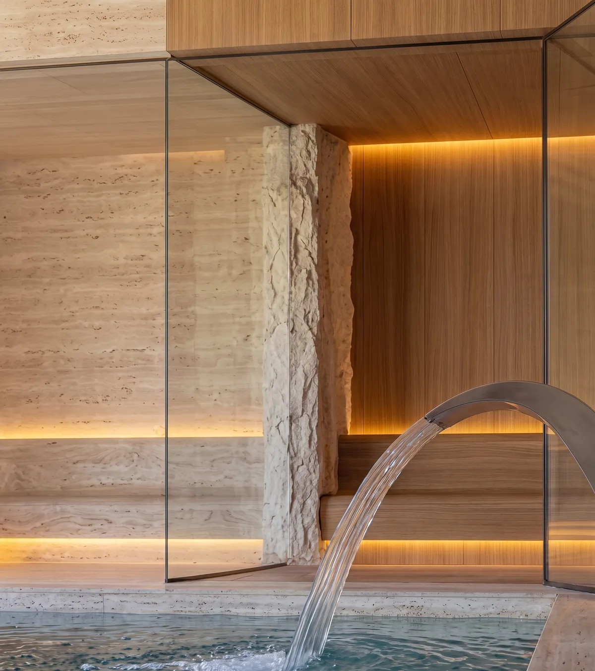 Detalhe da sauna e do spa da Casa Olive na Fazenda da Grama, com travertino iluminado, madeira e cascata sobre a piscina interna